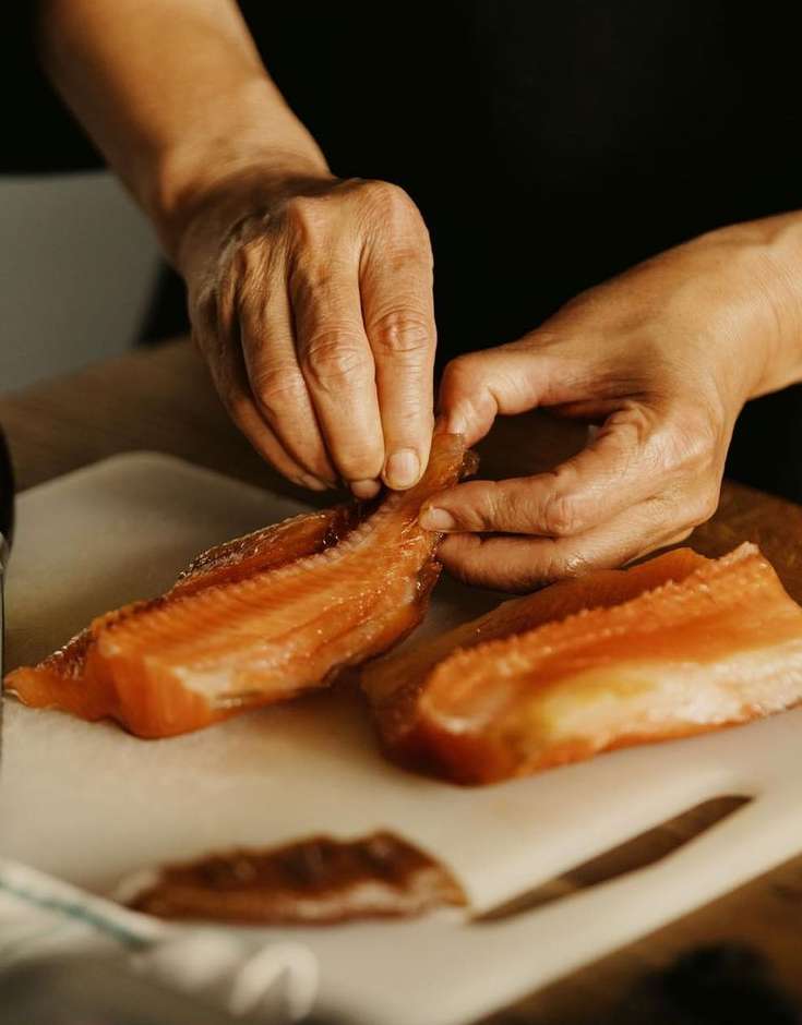 Close-up of an adult preparing salmon on a white cutting board, featuring the person's hands and orange-colored fish fillets indoors.