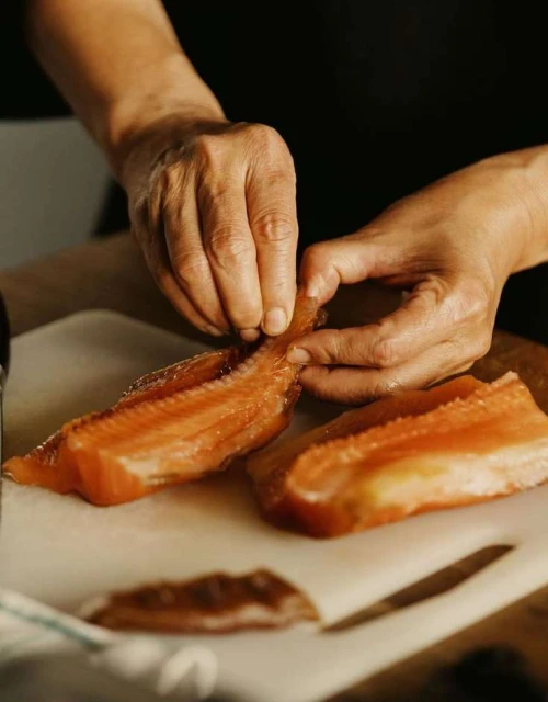 Close-up of an adult preparing salmon on a white cutting board, featuring the person's hands and orange-colored fish fillets indoors.