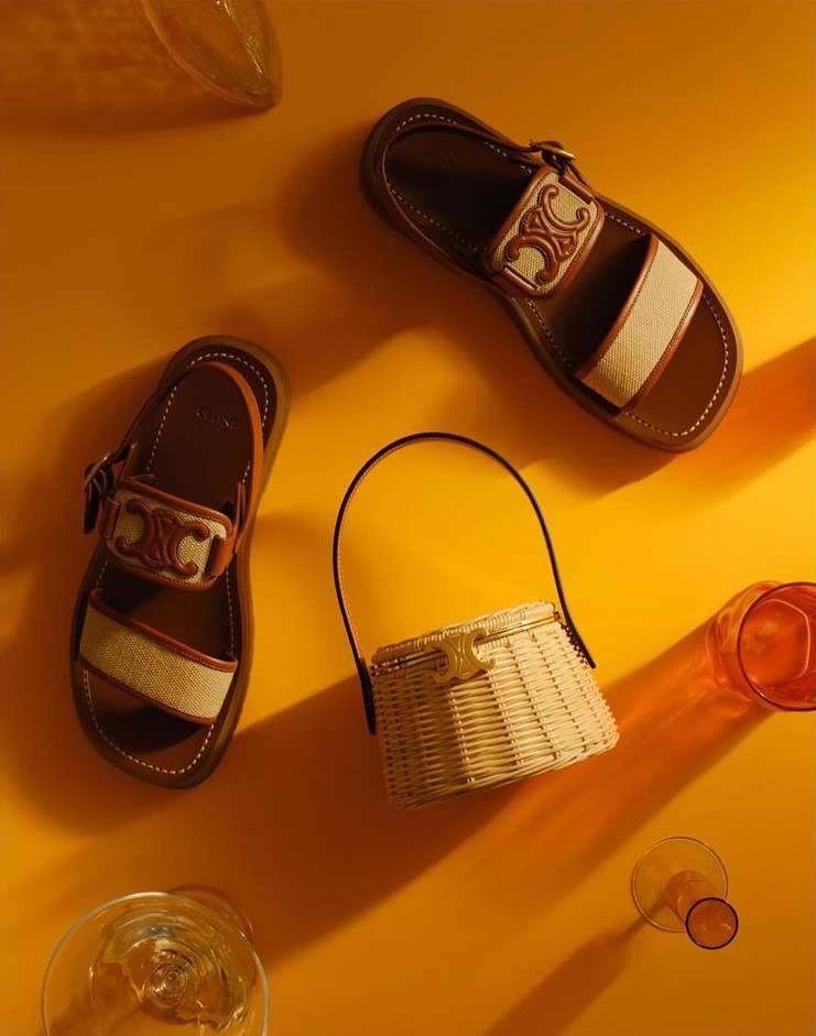 Flat lay of Celine leather platform sandals and straw handbag on a warm yellow background with clear decorative glass. Fashion product shot.