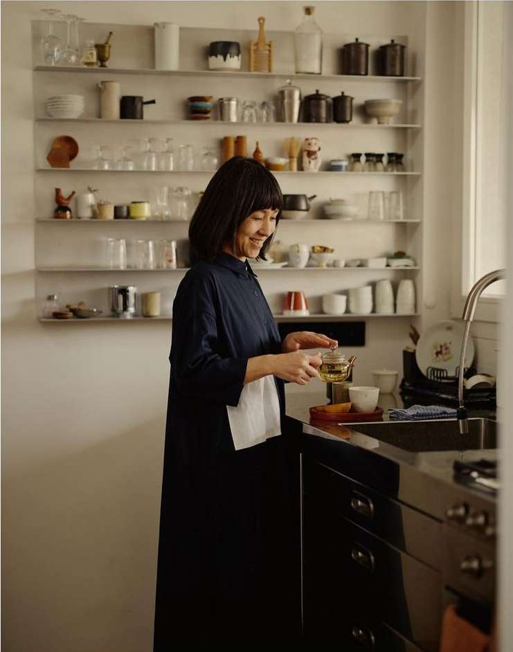 Asian woman in navy dress holding tea, smiling in kitchen with shelves of glassware and dinnerware, standing near dark cabinetry and a sink.