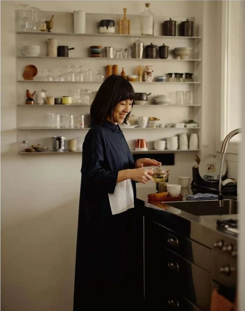 Asian woman in navy dress holding tea, smiling in kitchen with shelves of glassware and dinnerware, standing near dark cabinetry and a sink.
