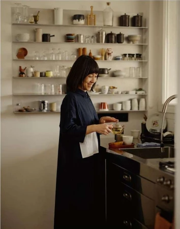Asian woman in navy dress holding tea, smiling in kitchen with shelves of glassware and dinnerware, standing near dark cabinetry and a sink.