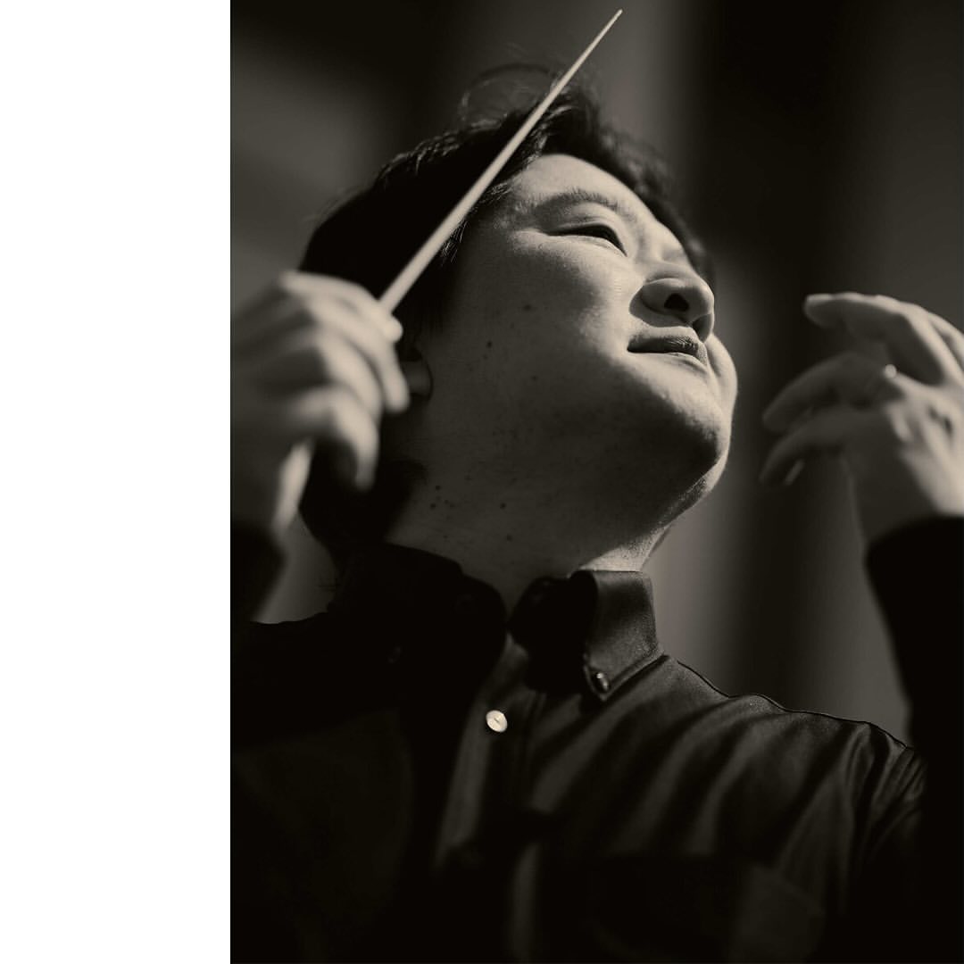 Asian man conducts music with baton in black and white portrait. Musical conductor in formal shirt leads orchestra, upward angle.