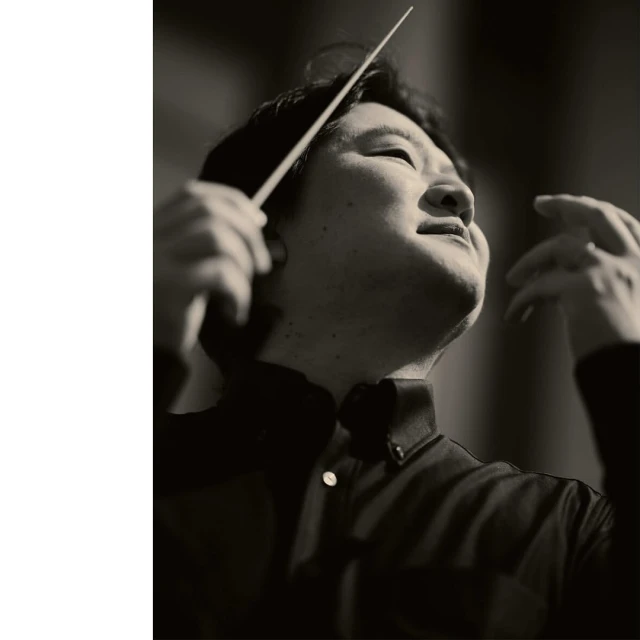 Asian man conducts music with baton in black and white portrait. Musical conductor in formal shirt leads orchestra, upward angle.