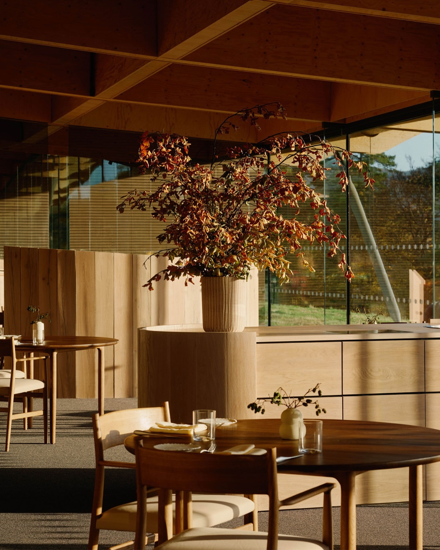 Interior of a restaurant with wooden tables and chairs, a large floral centerpiece, and natural light streaming in from large windows.
