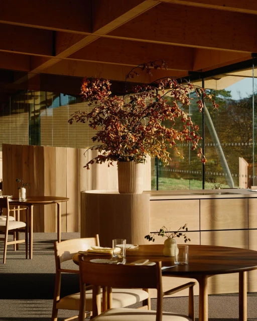 Interior of a restaurant with wooden tables and chairs, a large floral centerpiece, and natural light streaming in from large windows.
