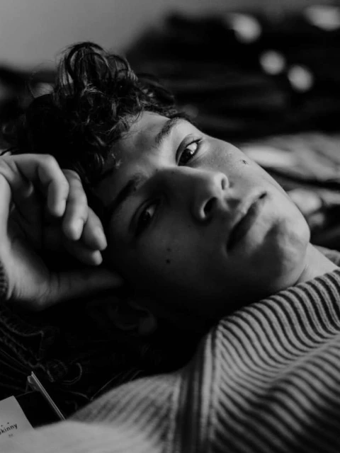 Black and white portrait of a young man with curly hair lying down and looking at the camera with a relaxed expression.