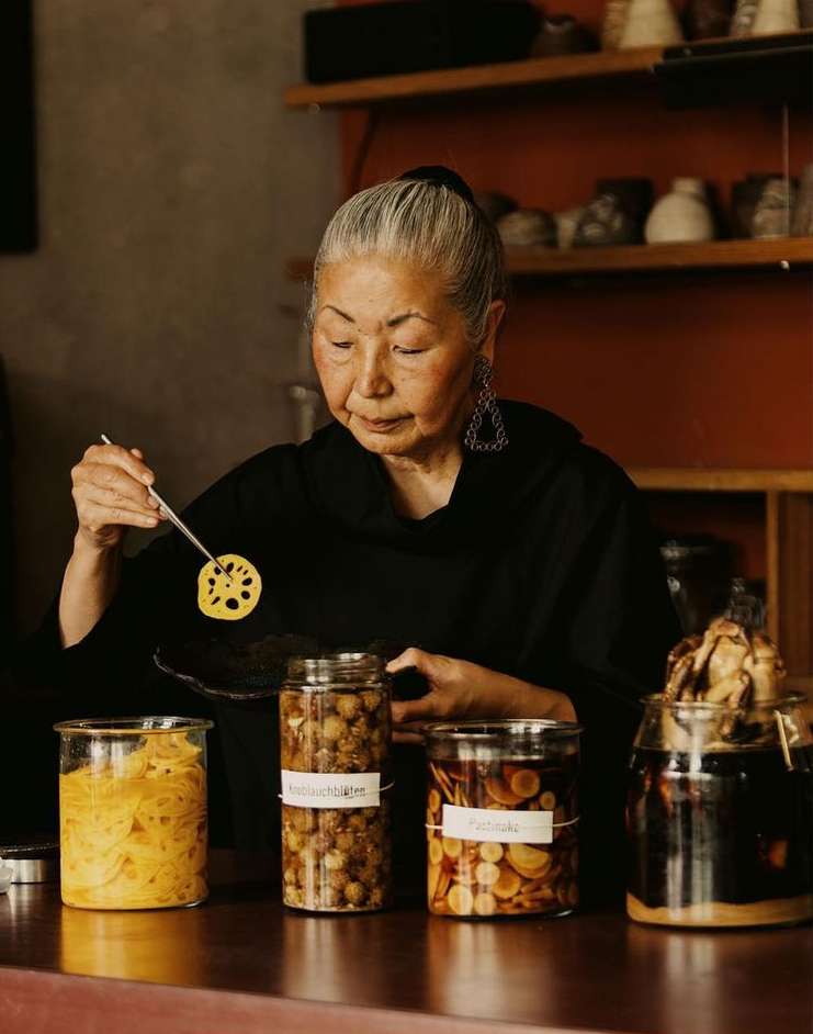 Older woman in black top using a small fork to inspect a sliced lotus root in a kitchen with glass jars of preserved foods.