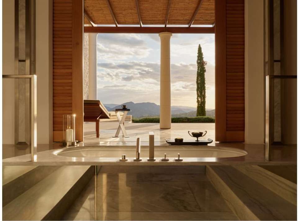 Luxury bathroom interior with a soaking tub in the foreground and an outdoor patio featuring a lounge chair and landscape in the background.