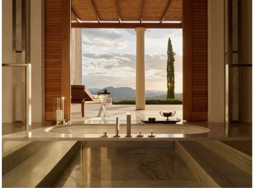 Luxury bathroom interior with a soaking tub in the foreground and an outdoor patio featuring a lounge chair and landscape in the background.
