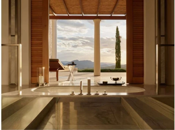 Luxury bathroom interior with a soaking tub in the foreground and an outdoor patio featuring a lounge chair and landscape in the background.