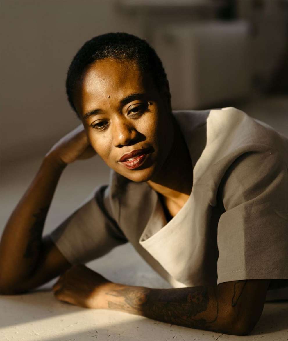 Indoor portrait of an adult woman with short hair lying down, with arm tattoo, wearing a grey blouse, and red lipstick on her lips.
