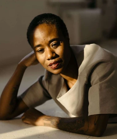Indoor portrait of an adult woman with short hair lying down, with arm tattoo, wearing a grey blouse, and red lipstick on her lips.