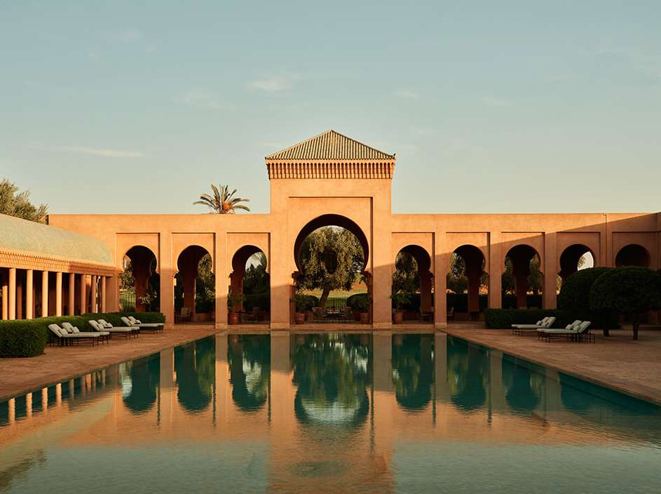 Exterior view of a luxury resort showcasing a tranquil swimming pool, terracotta-hued architecture with archways, and lush greenery, bathed in warm light.