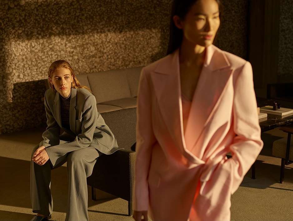 Two women in blazers pose indoors, one seated in grey and the other standing in pink, with a textured wall and soft, diffused lighting.