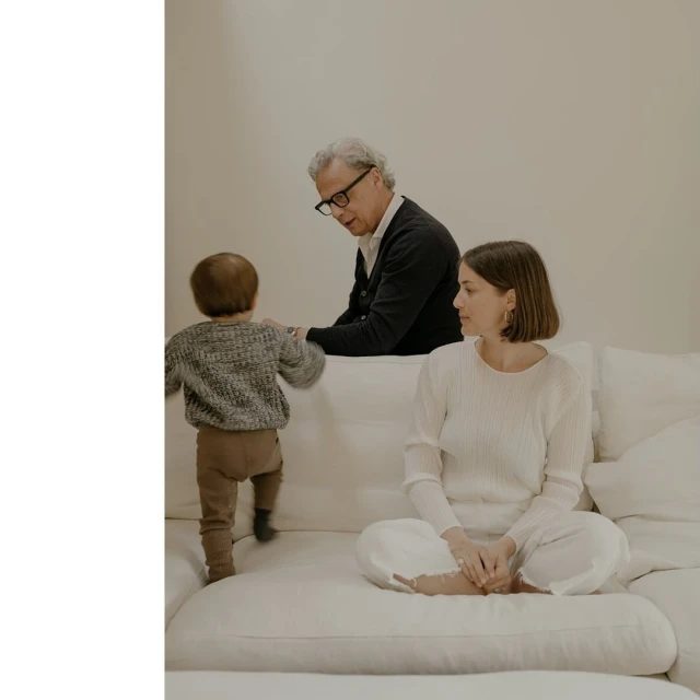 Family portrait indoors with a young child climbing onto a white sofa, an adult woman seated, and an elderly man observing.