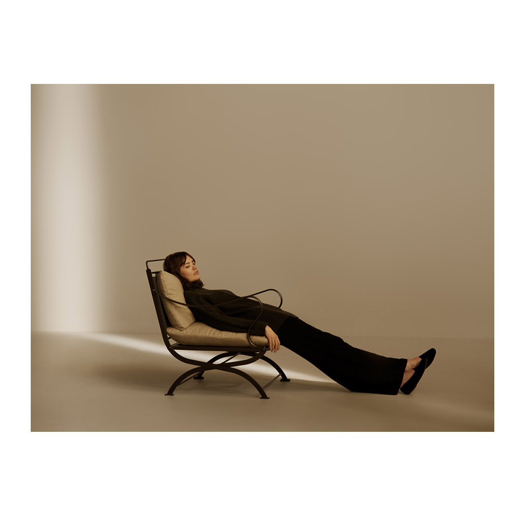 A woman wearing dark clothing is reclining in a chair in a relaxed pose with a neutral studio background and soft, diffused lighting.