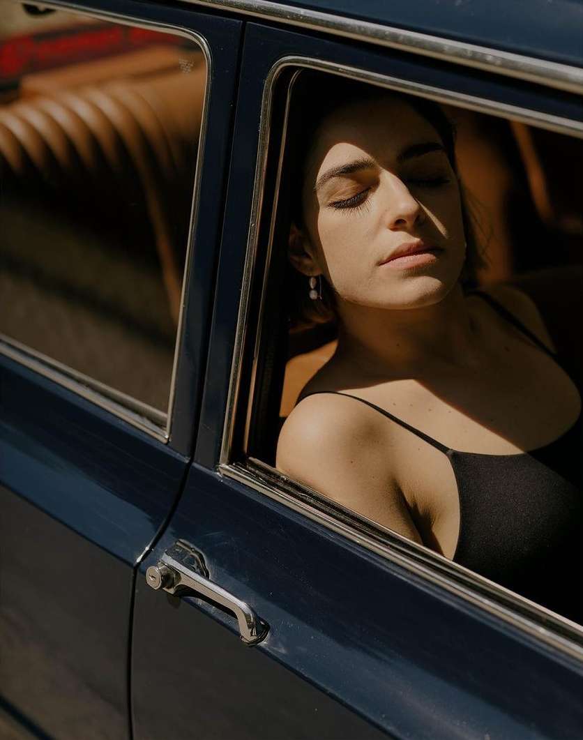 Woman with eyes closed sits in a blue car with brown interior. Light shines on her face as she rests. Silver car door handle visible.
