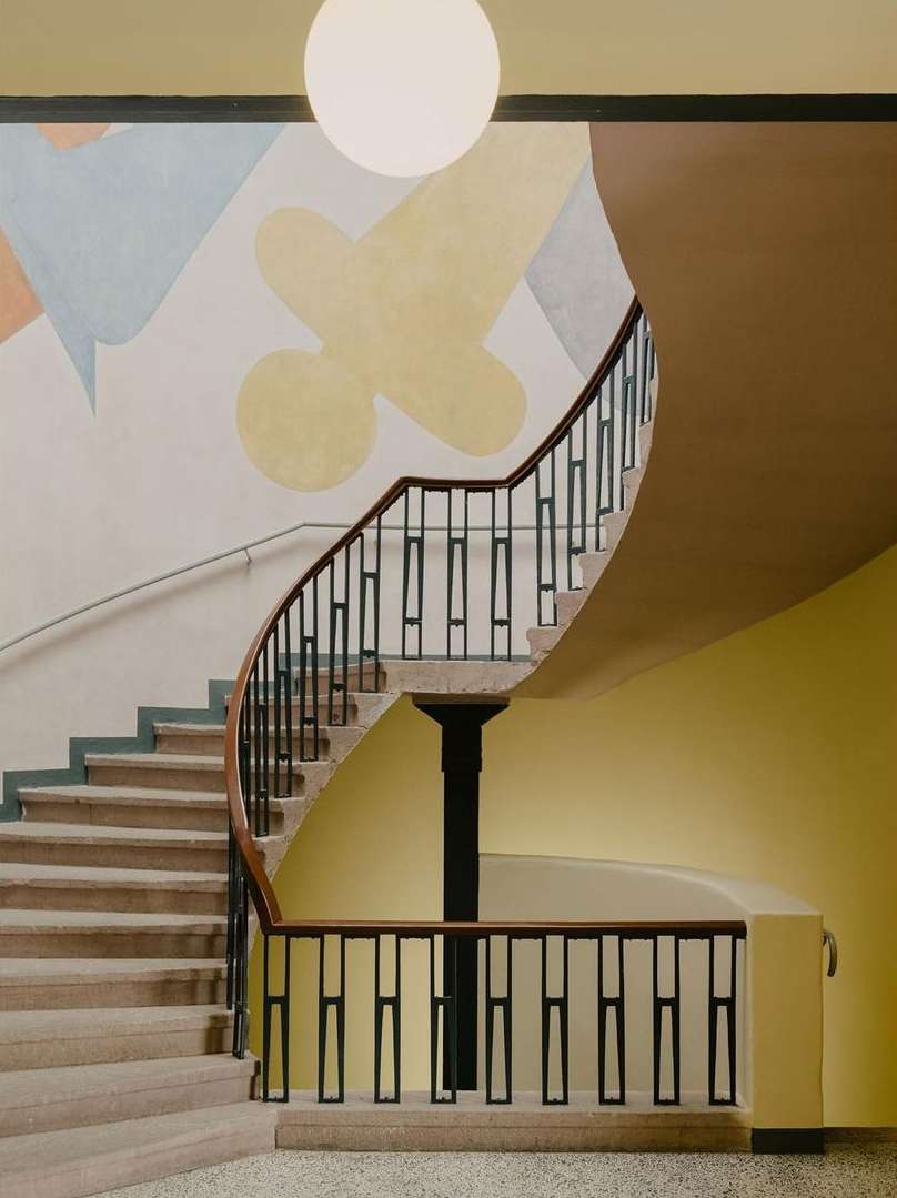 Interior view of a spiral staircase with light brown steps, a dark brown railing, a black support column, and a pastel abstract wall painting.