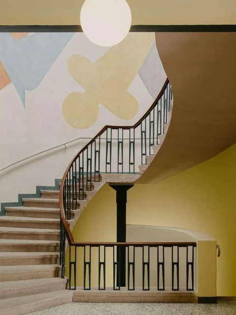 Interior view of a spiral staircase with light brown steps, a dark brown railing, a black support column, and a pastel abstract wall painting.