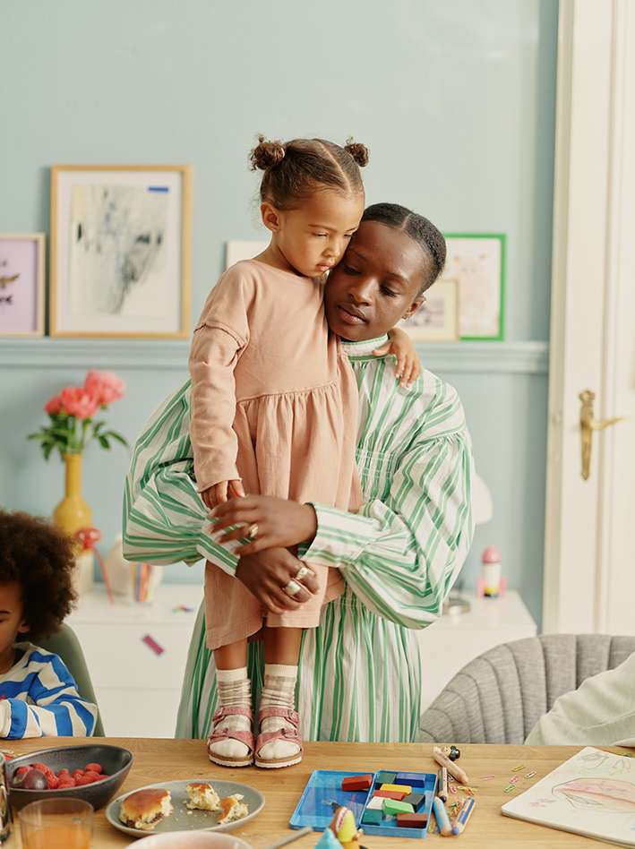An African-American mother holds her daughter at a table with breakfast and art supplies, in an indoor setting with soft, natural light.