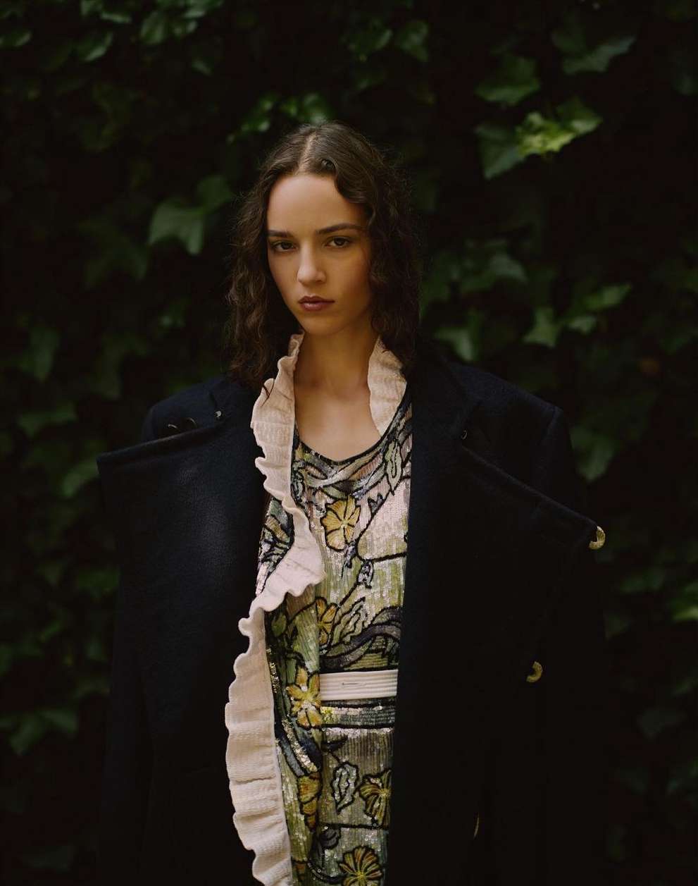 Portrait of a female model wearing a dark coat and floral dress outdoors against green foliage by Robert Rieger