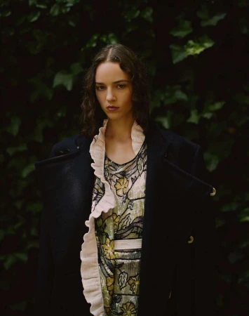 Portrait of a female model wearing a dark coat and floral dress outdoors against green foliage by Robert Rieger