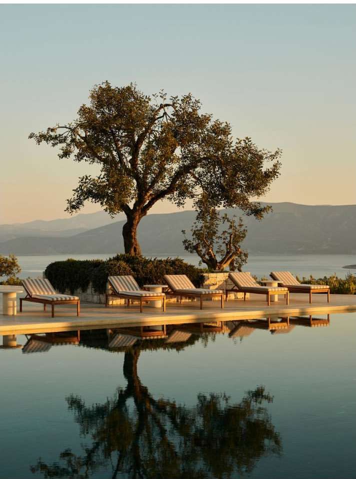 Tranquil outdoor scene featuring lounge chairs by an infinity pool reflecting a tree and distant mountains at golden hour.