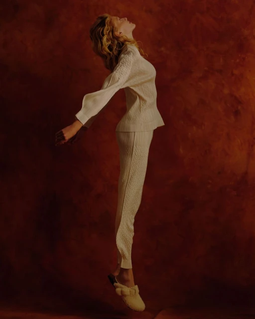 Woman in cream-colored pantsuit floating in studio with arms outstretched, looking up against a textured brown backdrop.
