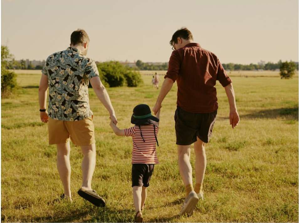 Two adult males hold hands with a child as they walk through a grassy field under a bright sky with distant trees and greenery.
