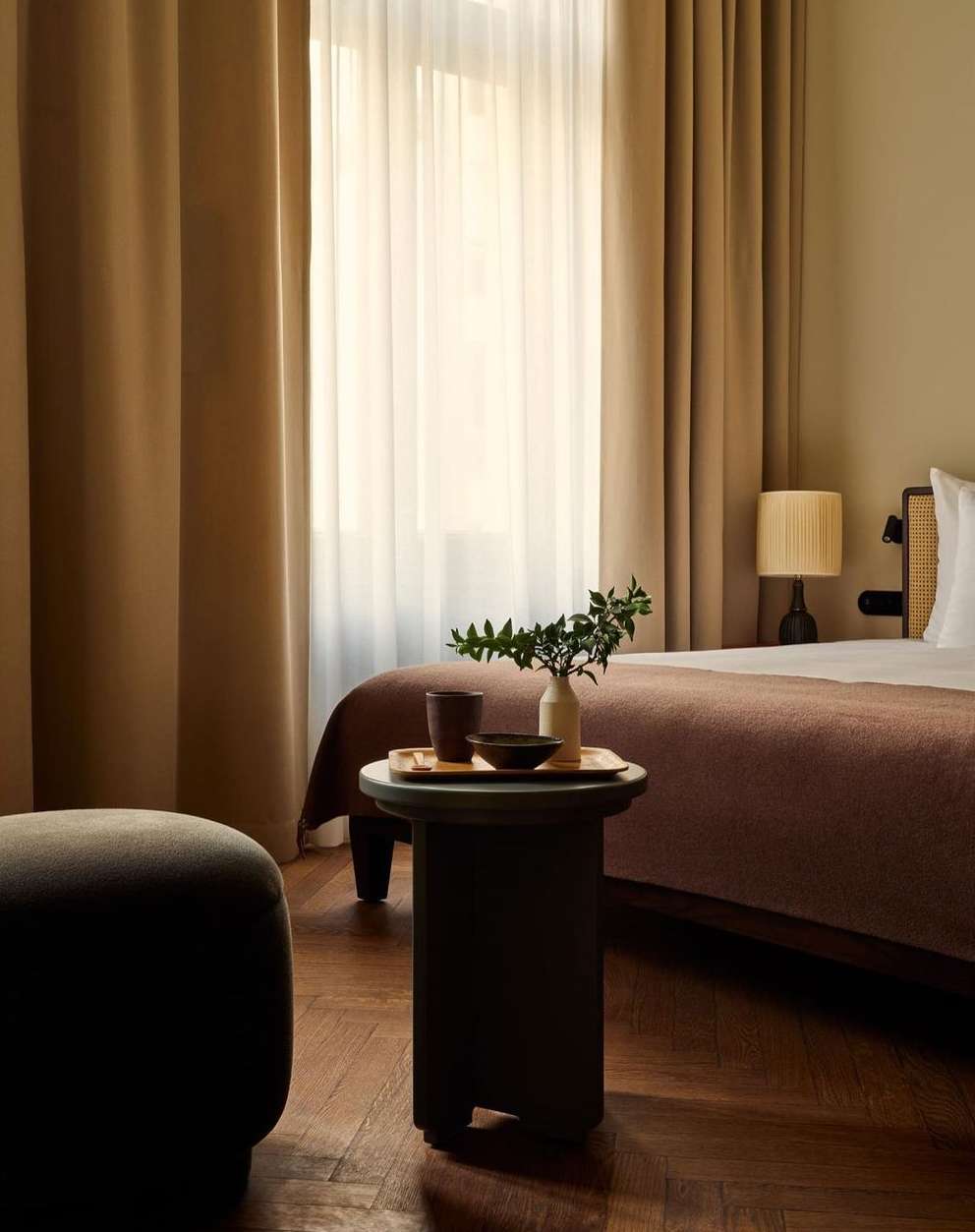 Bedroom interior featuring a small round table with a vase, cup, and bowl, set against a backdrop of beige curtains and bedding.