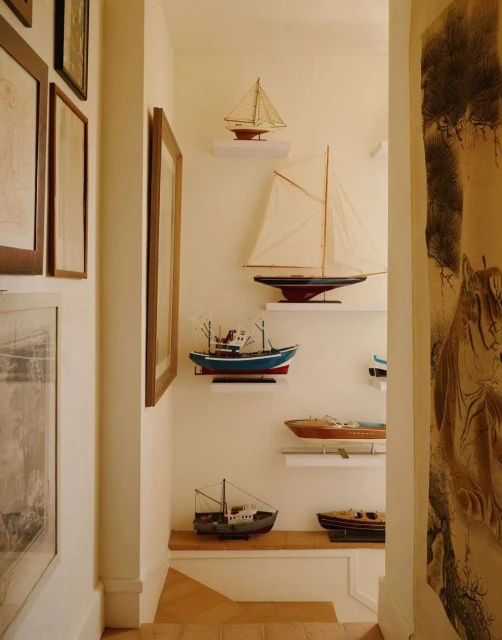 Collection of decorative model sailboats and speedboats displayed on shelves in a home interior with framed wall art.