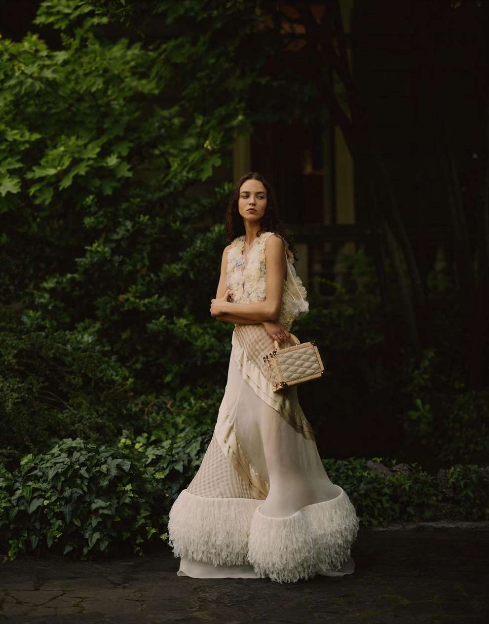 A solitary female fashion model poses in an ivory dress and vest with a handbag amidst a lush green garden setting, capturing outdoor elegance.