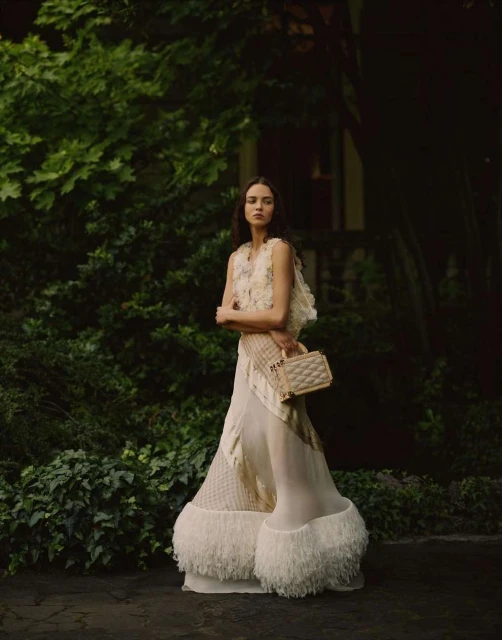 A solitary female fashion model poses in an ivory dress and vest with a handbag amidst a lush green garden setting, capturing outdoor elegance.