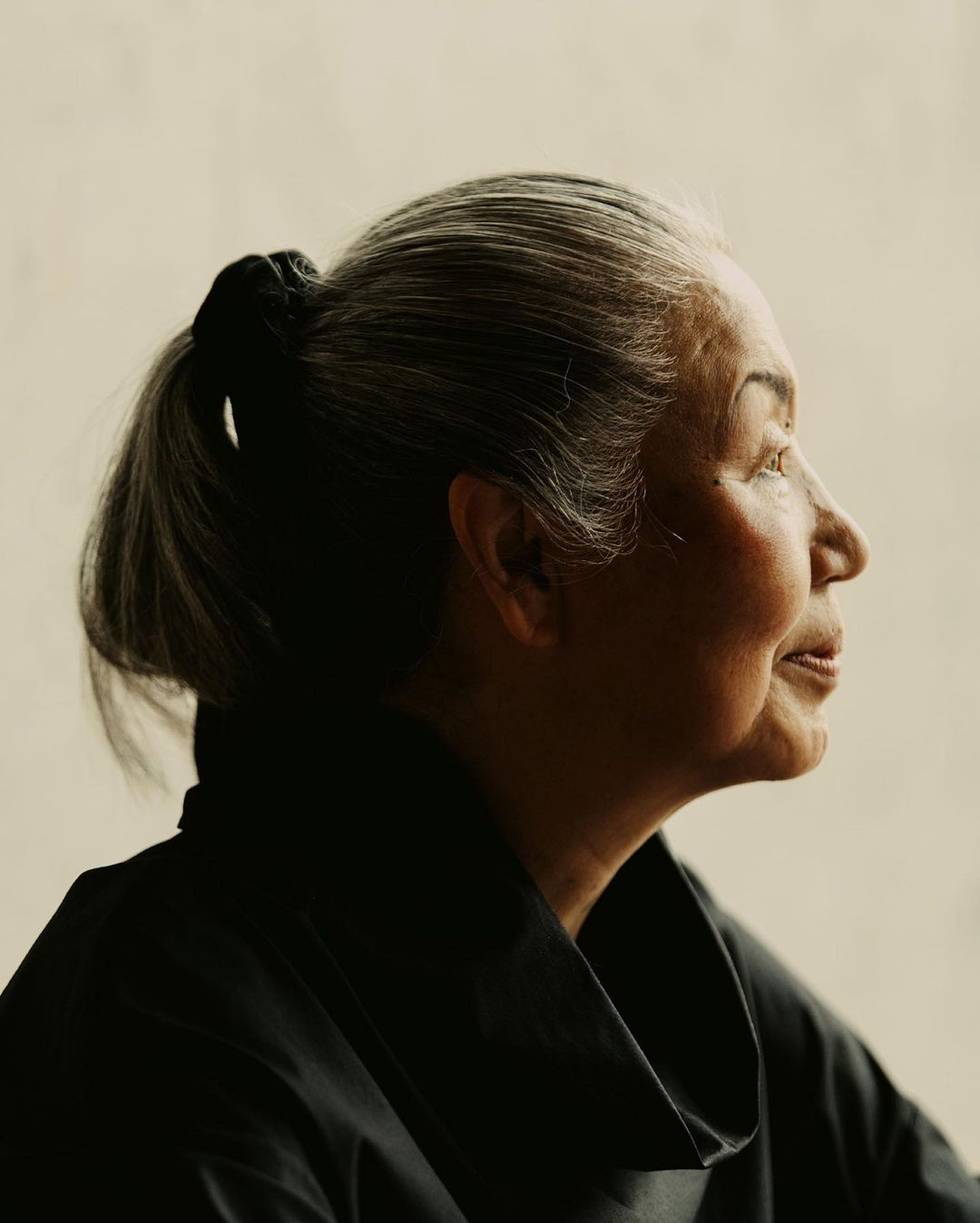 Profile portrait of a mature woman with gray hair in a ponytail wearing a black top, set against a light neutral background indoors.