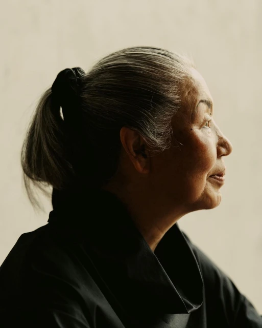 Profile portrait of a mature woman with gray hair in a ponytail wearing a black top, set against a light neutral background indoors.