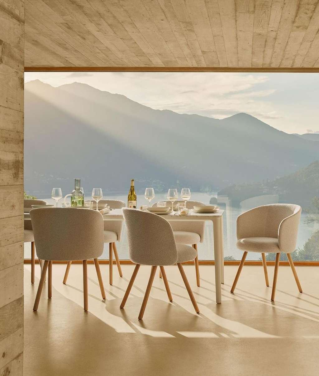 Dining room interior with a white table set with plates and glassware surrounded by upholstered chairs, a scenic view through a window.