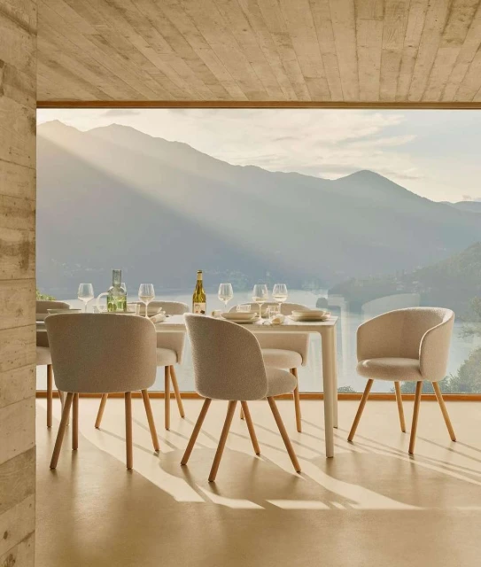 Dining room interior with a white table set with plates and glassware surrounded by upholstered chairs, a scenic view through a window.