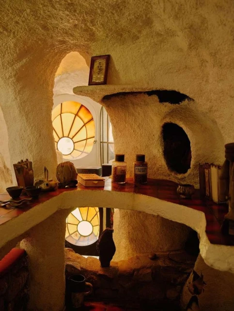 Interior view of a uniquely designed room with textured walls, stained glass window, shelves displaying decorative objects and containers.