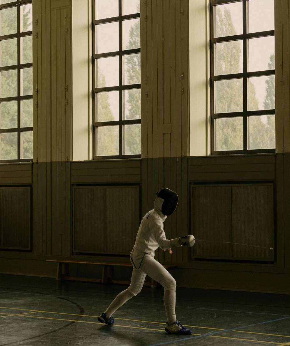 An indoor shot of a fencer in protective gear in action on a dark surface inside a gymnasium, near large windows, with trees visible.