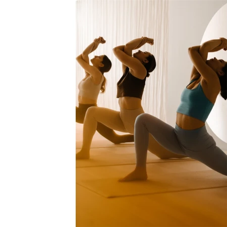Three adult females are in a yoga studio doing a modified warrior pose with soft, diffused lighting, wearing athletic wear on yoga mats.