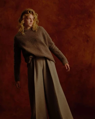 Portrait of a woman with curly blonde hair wearing a brown knitted sweater and wide-leg trousers against a textured rust-red background.