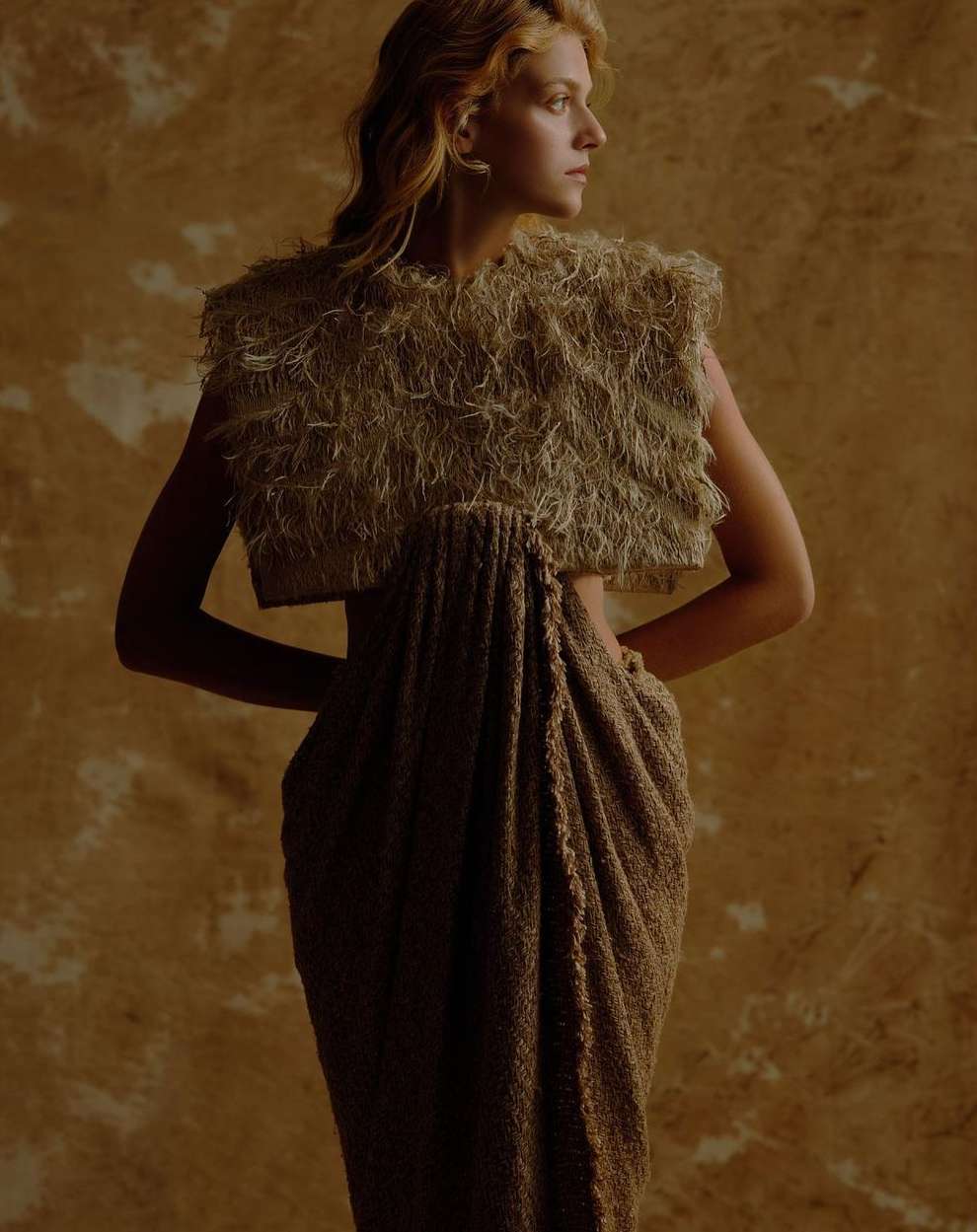 A fair-skinned woman with wavy blonde hair is wearing a beige textured top and draped skirt against a mottled tan background.