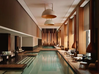 An indoor swimming pool hall in Berlin featuring warm wood paneling, large dome pendant lights, and arranged lounge seating along the edge.