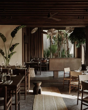 Interior view of a restaurant with wooden tables, woven chairs, tropical plants, and a ceiling fan photographed by Robert Rieger
