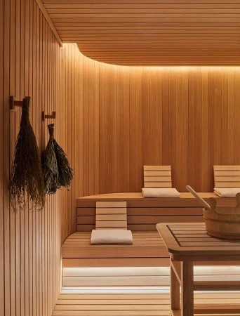 Modern wooden sauna interior with slatted walls, tiered benches, white cushions, and hanging birch whisks.