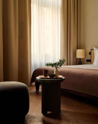 Minimalist bedroom interior with a round side table, a wooden tray, and sunlight streaming through sheer curtains