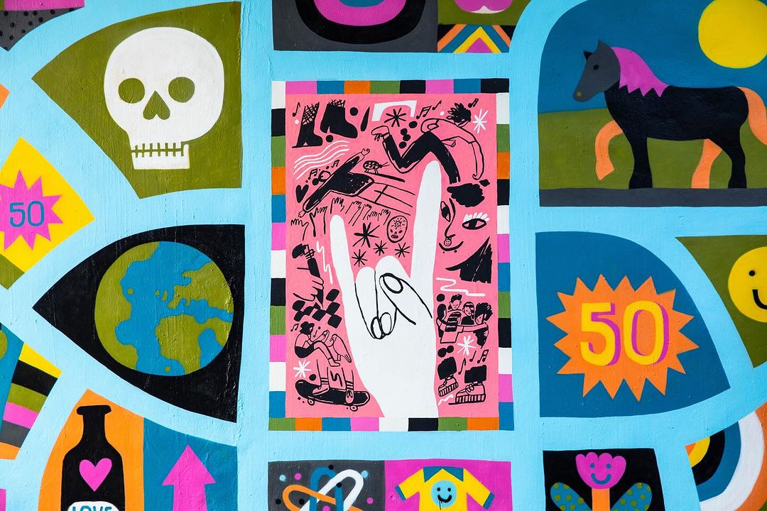 Vibrant and colorful mural with diverse imagery, including a skull, earth, hand gesture, horse, flower, and abstract shapes on a light blue background.