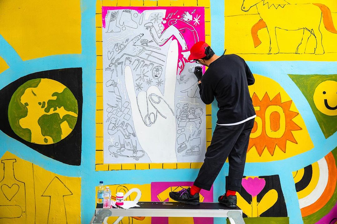 An artist in a red cap paints a mural with a large hand gesture, set against a colorful backdrop of yellow, blue, and white.