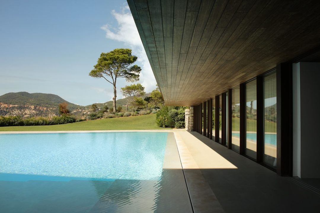 Modern architecture featuring a turquoise swimming pool, manicured lawn, and a backdrop of lush green hills against a blue sky.